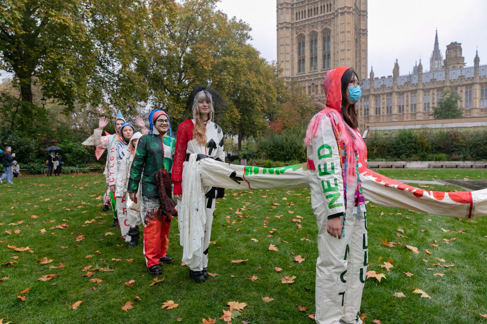 A line of people in uniquely-customised suits connected together, standing in a park.