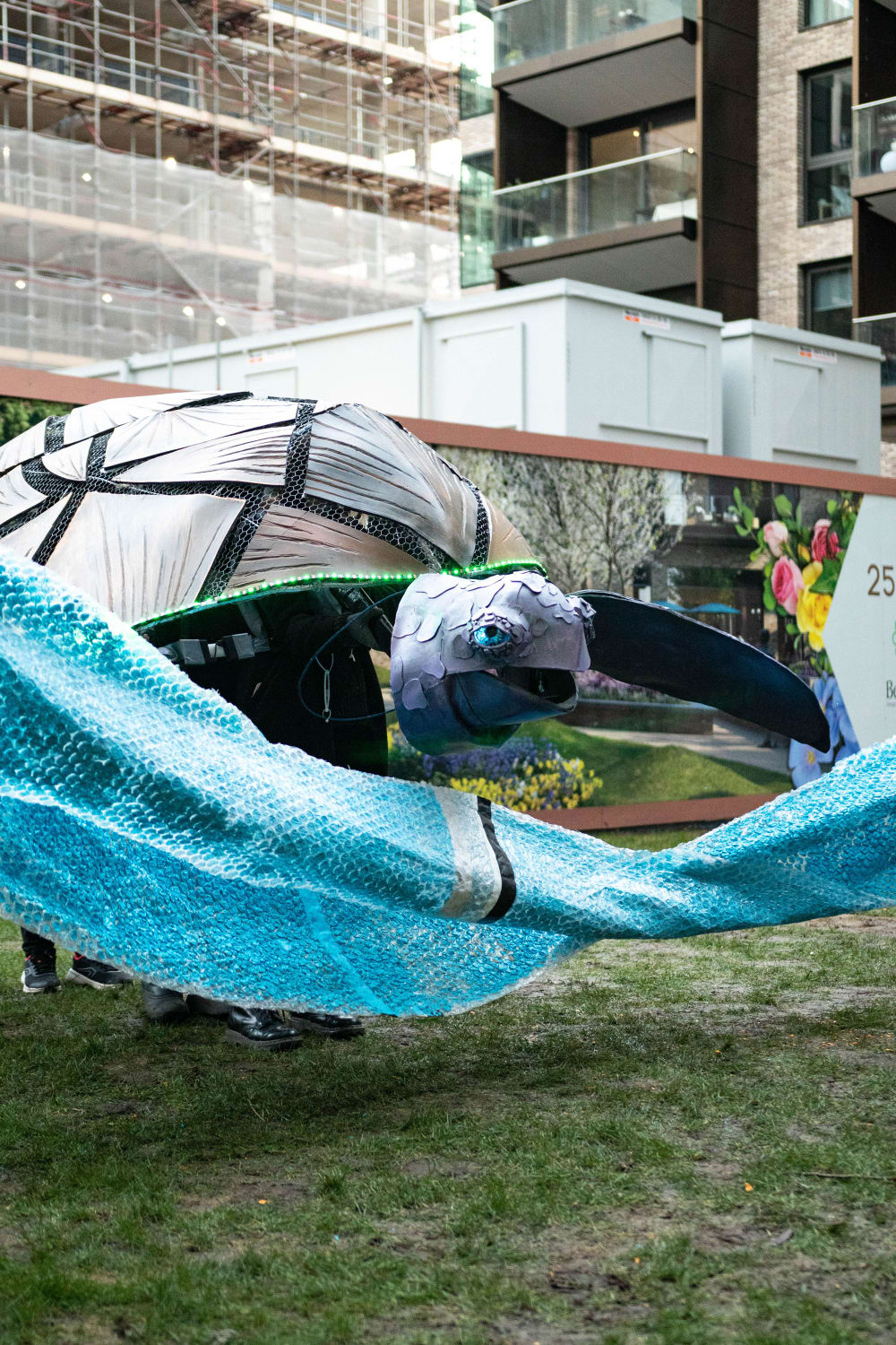 A large puppet of a turtle with geometric lines on its shell and blue eyes is operated behind a swathe of blue bubblewrap material in an outdoor setting with backdrop of scaffolding.