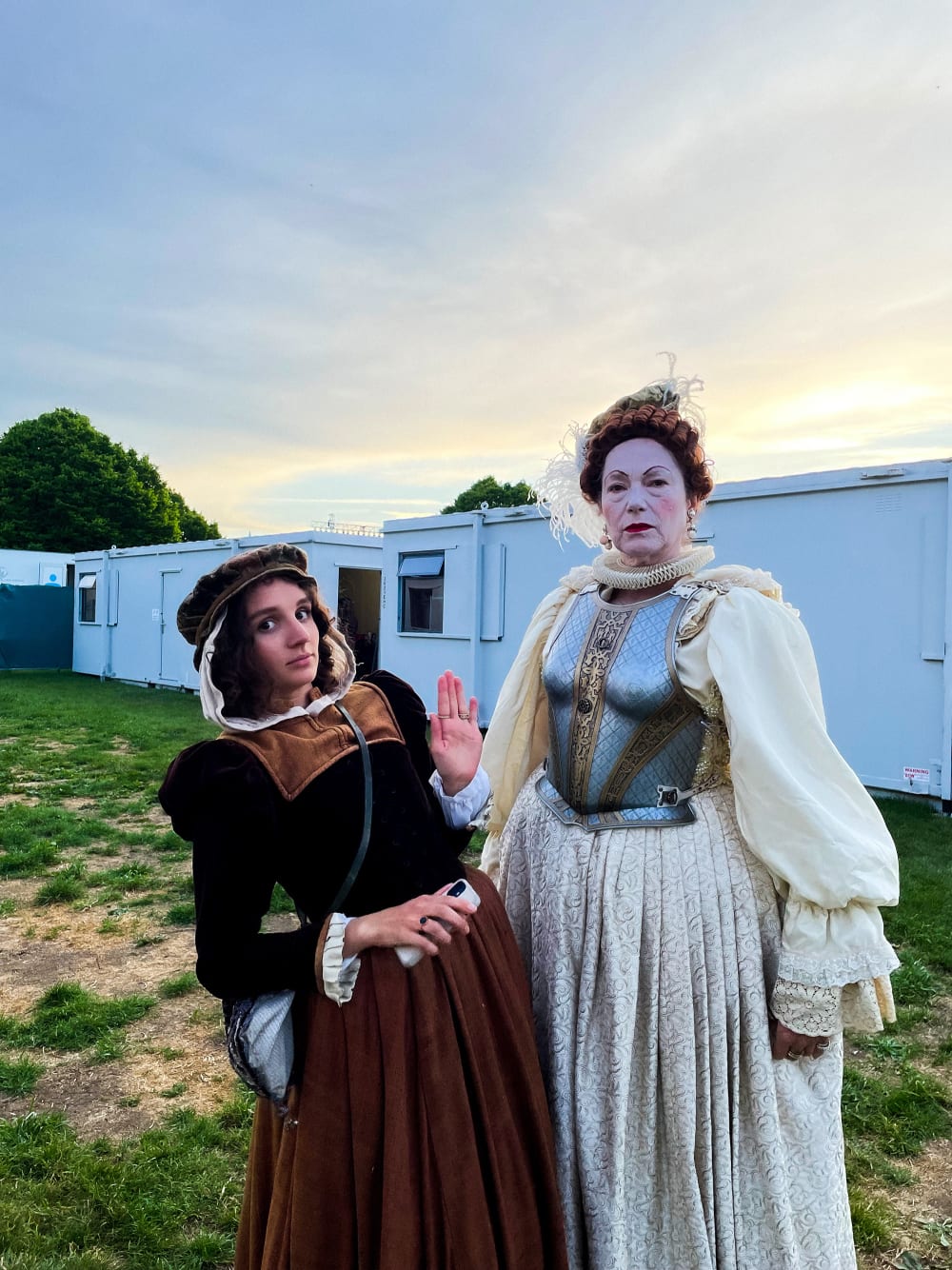 Two performers pose in Elizabethan costumes on grass among trailers  backstage 
