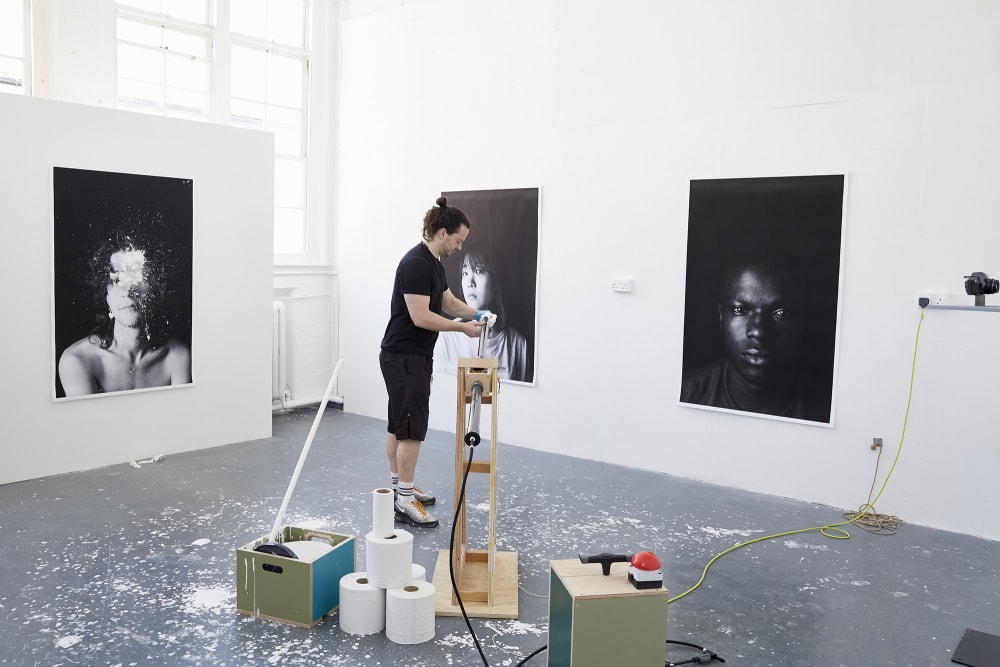 The artist handles his installation piece, an interactive air pump cannon which is used to fire paint at the portraits. 3 large scale black and white portraits surround him.