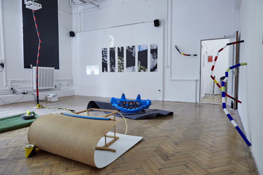 A room shot of The Postgraduate Fine Art Show 2022. Installation pieces by MA Fine Art: Drawing students. Includes a structure of curved wooden board and yellow and black tape. There are also installation pieces attached to the walls, such as colourful tubing.