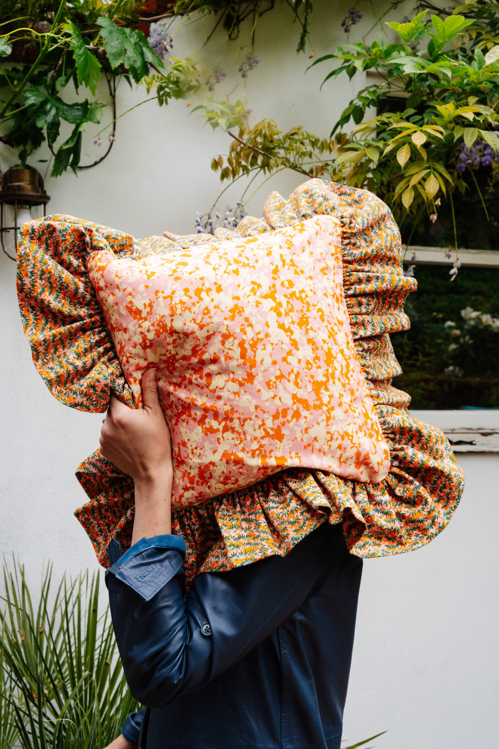 A person stood outside holding a cushion over their face. The material of the cushion is pink and orange pattern; the large frill around the outside is orange and green