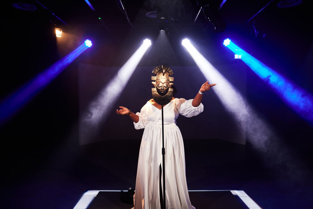 A shot of a performance, a woman in a long white dress is standing before a mask on a stand