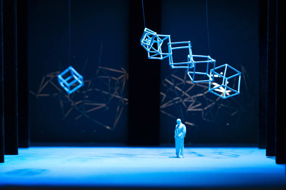 A miniature model installation with hollow cube structures hanging from the ceiling, there is a small blue man standing underneath them