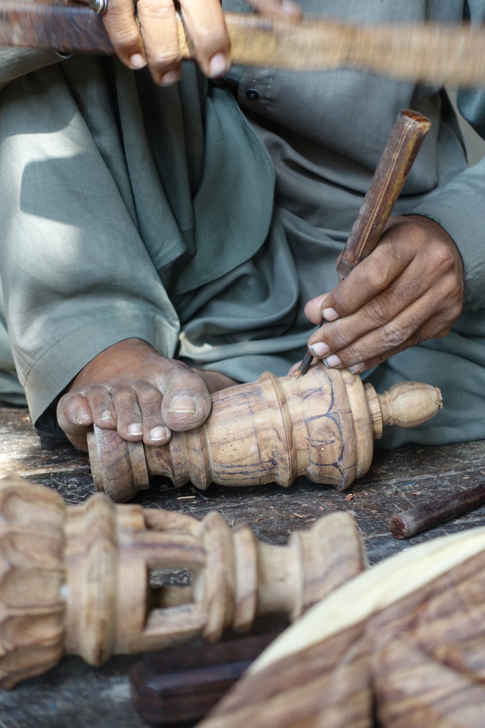 A close up picture of a person intricately carving a piece of wood. They are holding a wood carving tool in each hand and holding the piece of wood in place with their feet.  