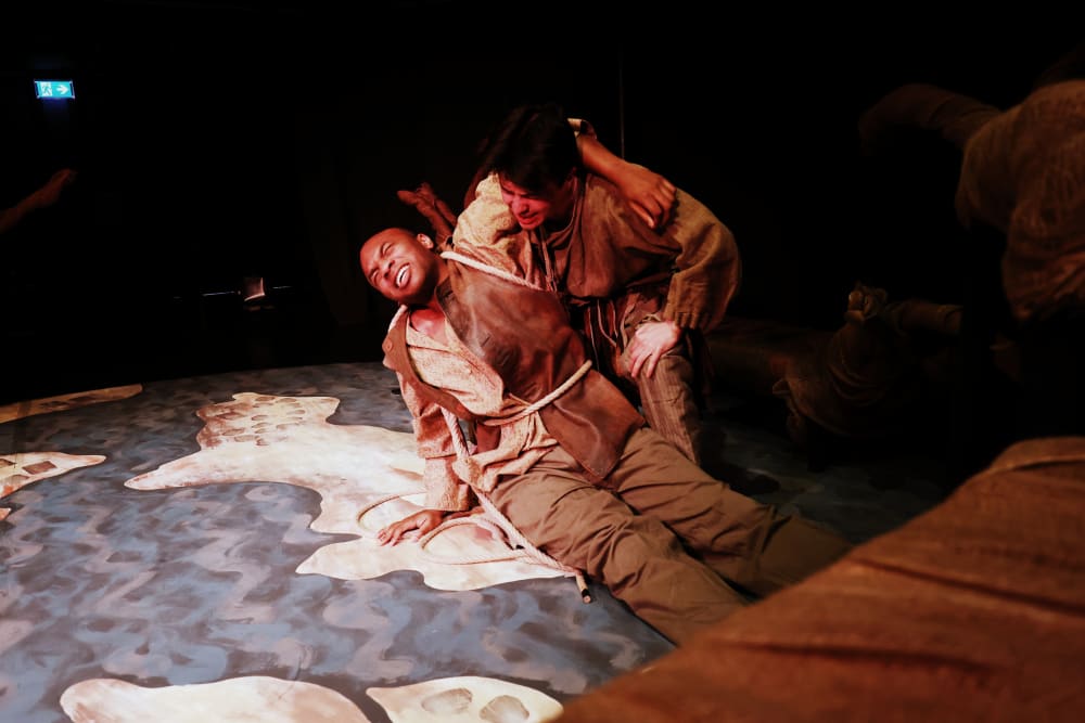 Image shows a person lying down, apparently in pain, while another person holds them. They are wearing brown clothing with ropes wrapping round them both.