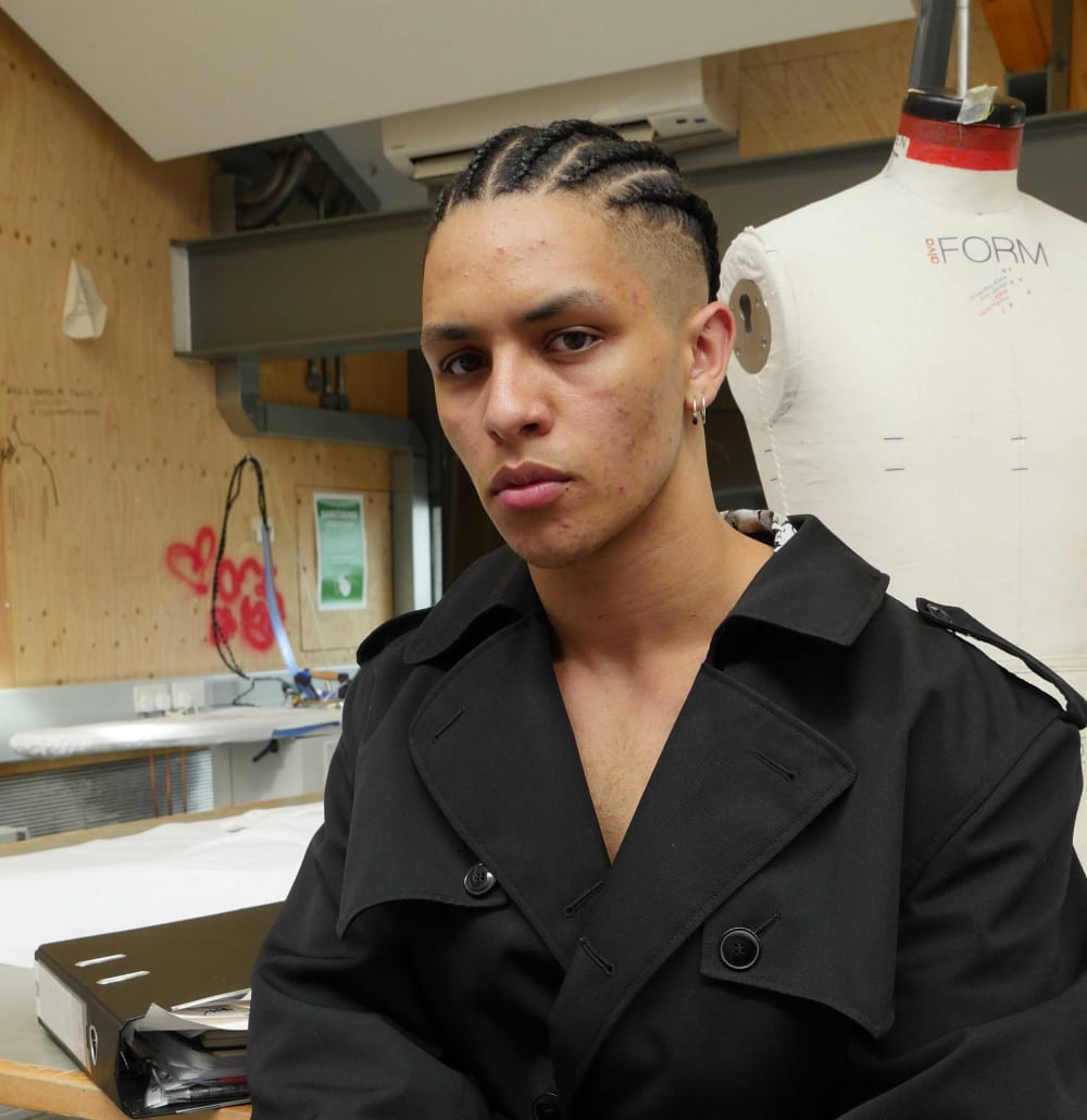 A young man in a long black coat with his hands in his pockets. 