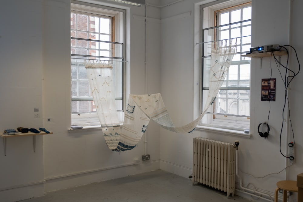 Image shows a long piece of embroidered fabric draped between 2 windows in the corner of a white room.