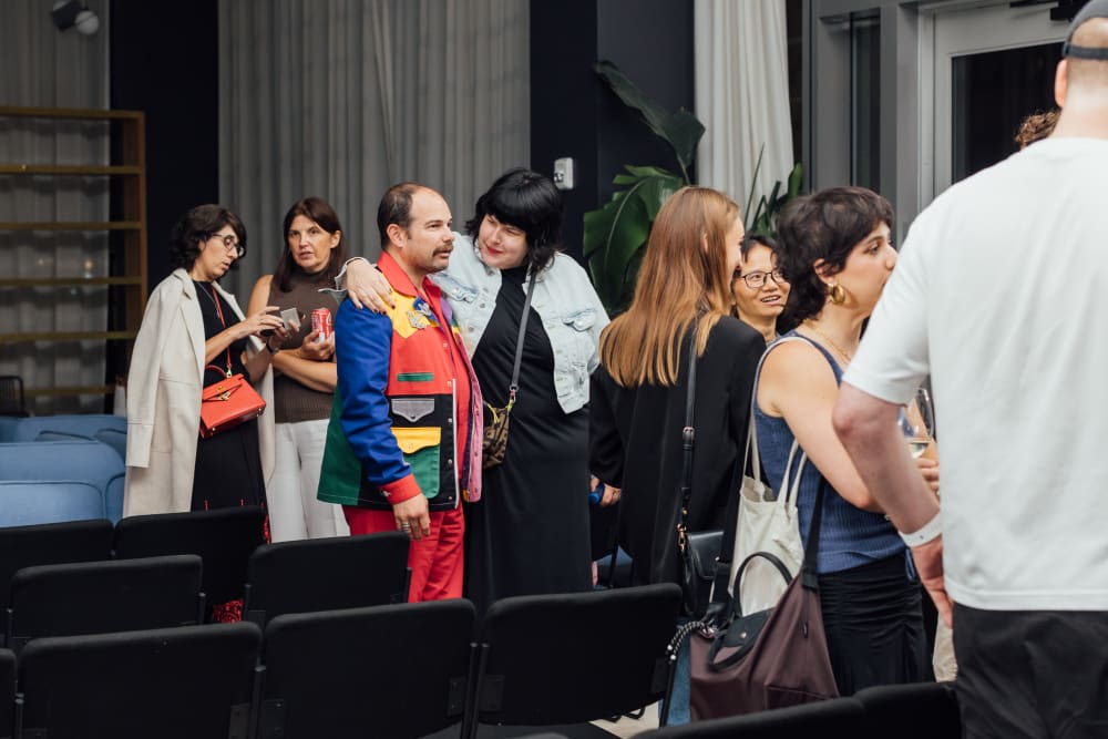 Guests standing amongst the seating, chatting. A woman has one arm around a man's shoulders. 