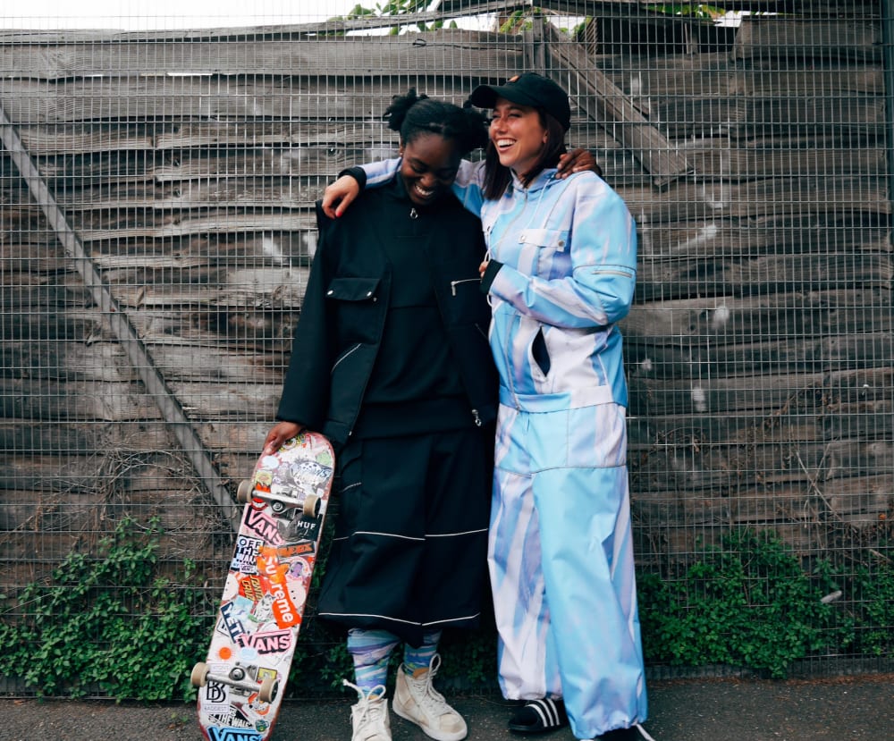 Two female skateboarders in reflective sportswear, one with the arm around the other.