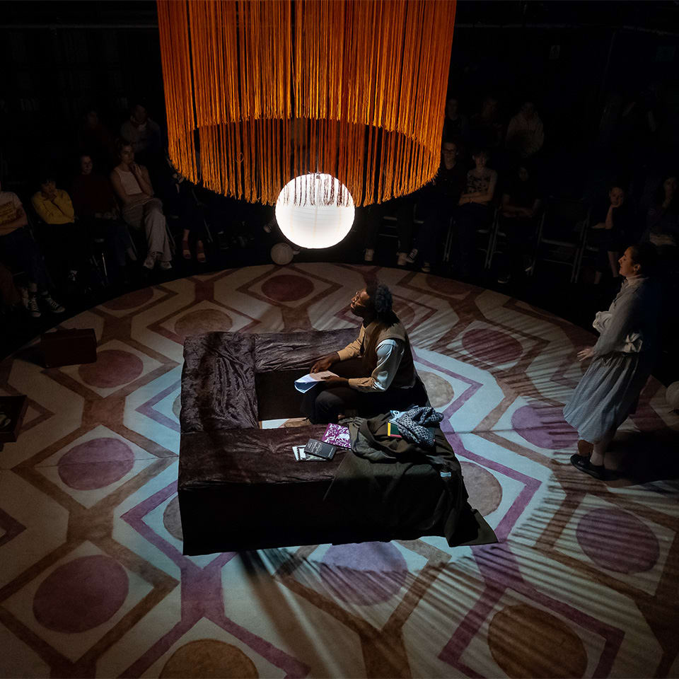 An actor is seated in the middle of a circular stage surrounded by an audience, beneath an orange fringe lamp. The stage floor has a retro geometric pattern.