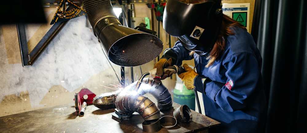 A young woman wearing protective gloves and working on a complex metal sculpture made of curved and tubular steel pieces. 