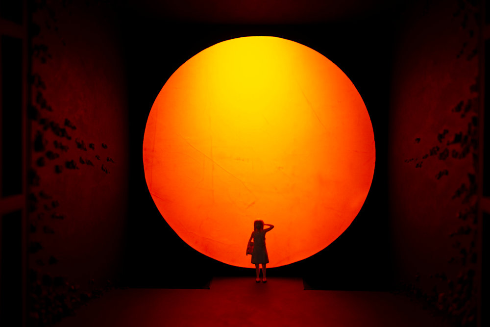 Silhouette of a child standing before a massive glowing orange and yellow circular light installation in a dark immersive space, creating a dramatic futuristic and surreal atmosphere.