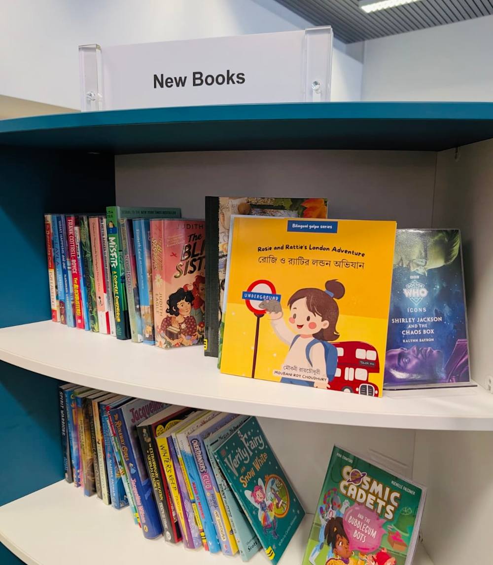 A bilingual Bengali-English children’s book, Rosie and Rattie’s London Adventure, displayed on a library shelf under a “New Books” sign.