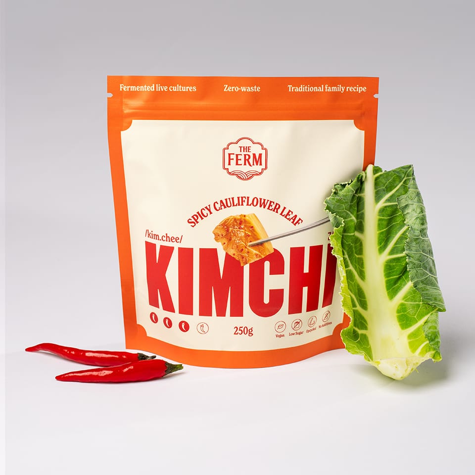 An orange pouch of The Ferm's spicy cauliflower leaf kimchi next to a green cauliflower leaf.