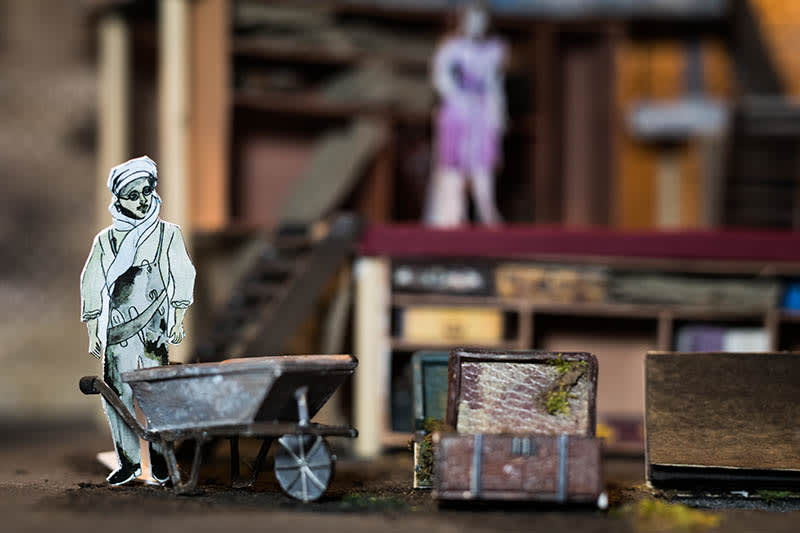 A small scale set design of a man with a wheelbarrow and suitcase