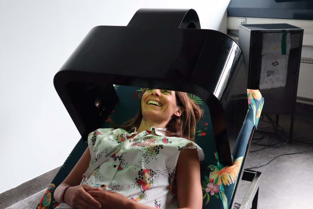 A woman lies smiling in strange contraption with black screen tilted at an angle over her face.