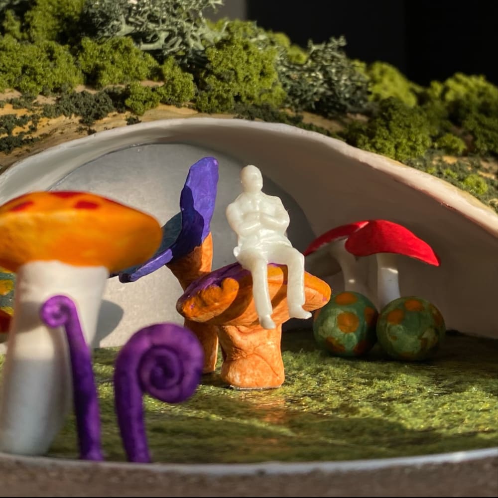 The focus is on a model of a person, who is sat on a purple and orange mushroom, there are other smaller mushrooms surrounding them 