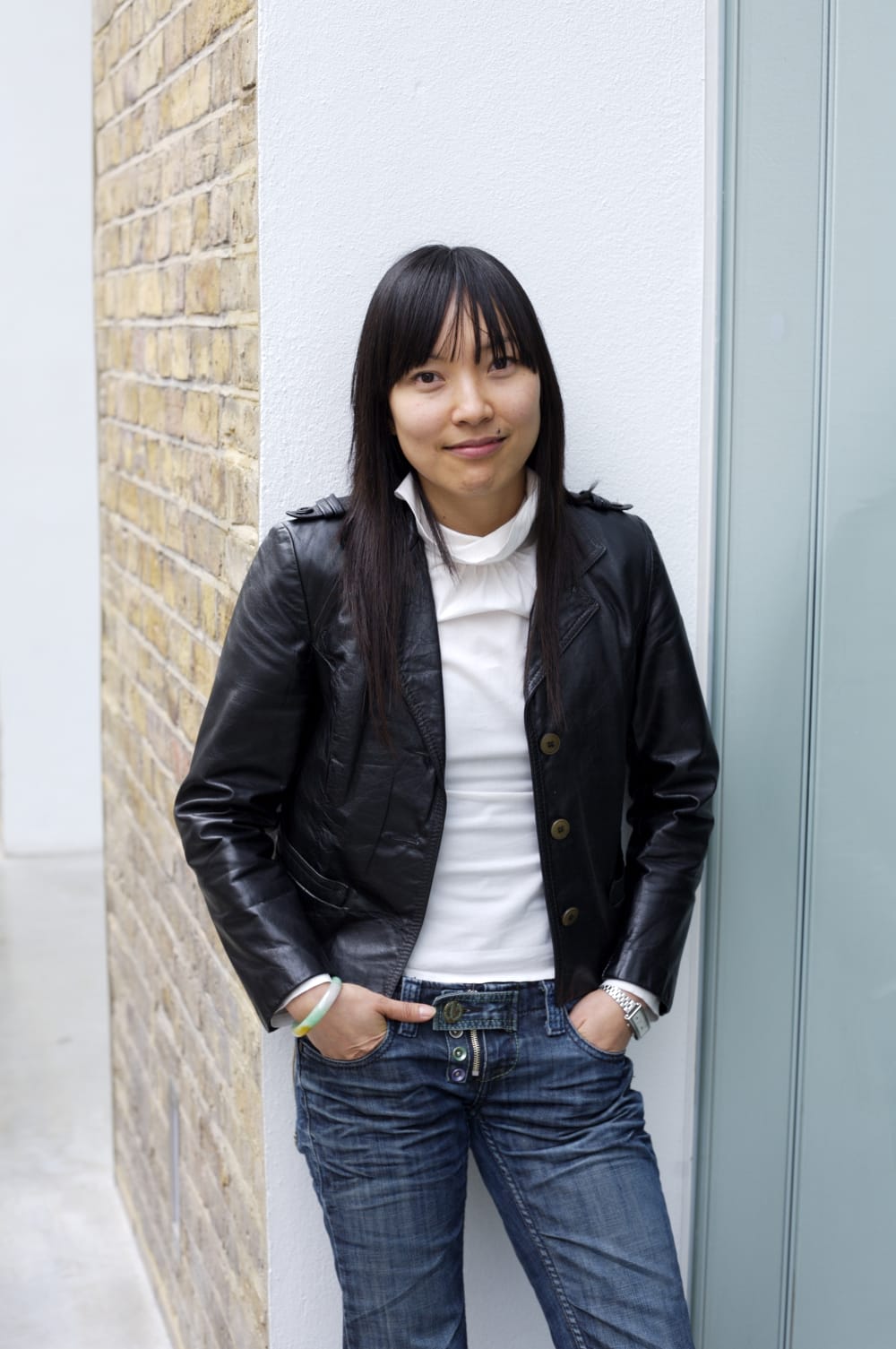 Portrait photo of Suki stood against a a pale background. She's wearing blue jeans, a black jacket and white t-shirt, her hands are in her pockets and she's smiling into the camera