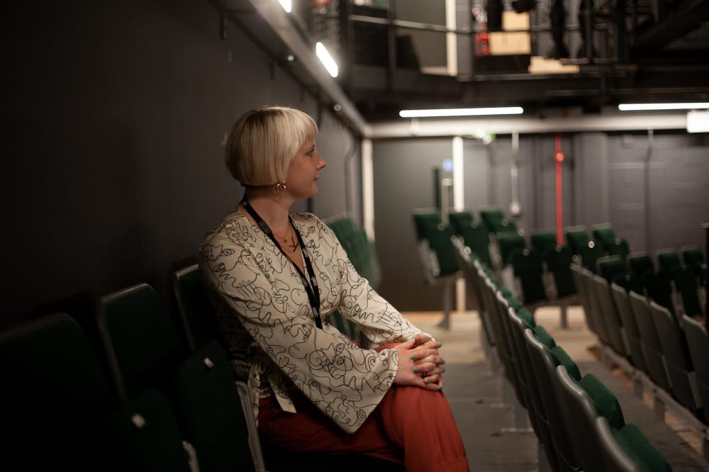 A person sits in an empty theatre and amongst red seats. She looks towards the stage which is out of frame.