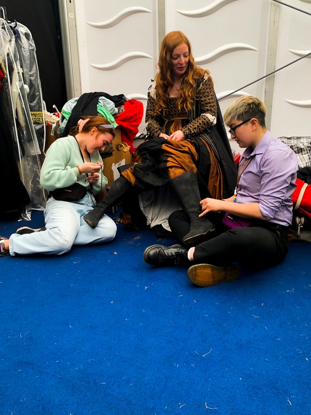 Two costume designers kneel on the ground while working on the boots of a performer who is wearing the boots and chatting to them    
