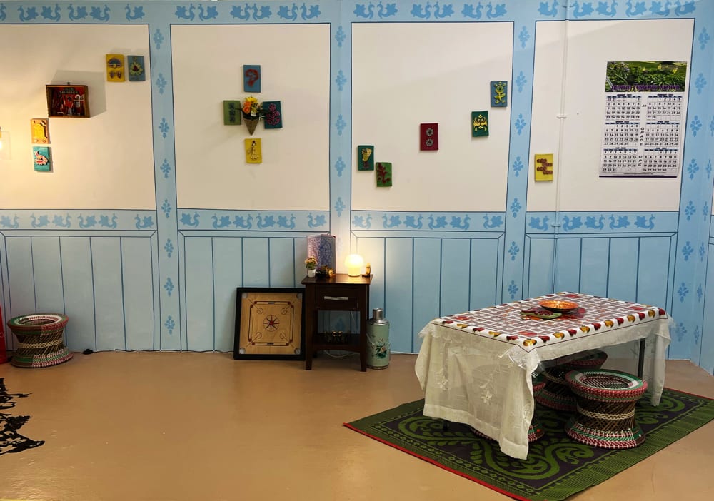 An installation resembling a domestic space. There is a table with a tablecloth, and stool positioned on top of a rug. Shapes made from tea stencils are on the floor. The walls are decoratively painted with blue and white and a number of small ceramic squares with designs on hangers on the walls.  