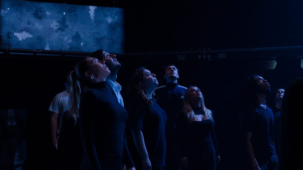Image shows a dark stage with several people dressed in black looking up towards a dim light.