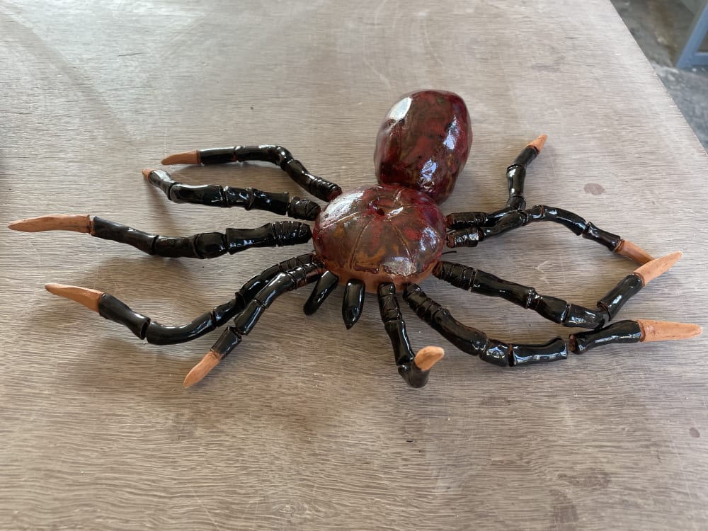 Photograph of a red and black glazed terracotta spider sculpture on a wooden table. 