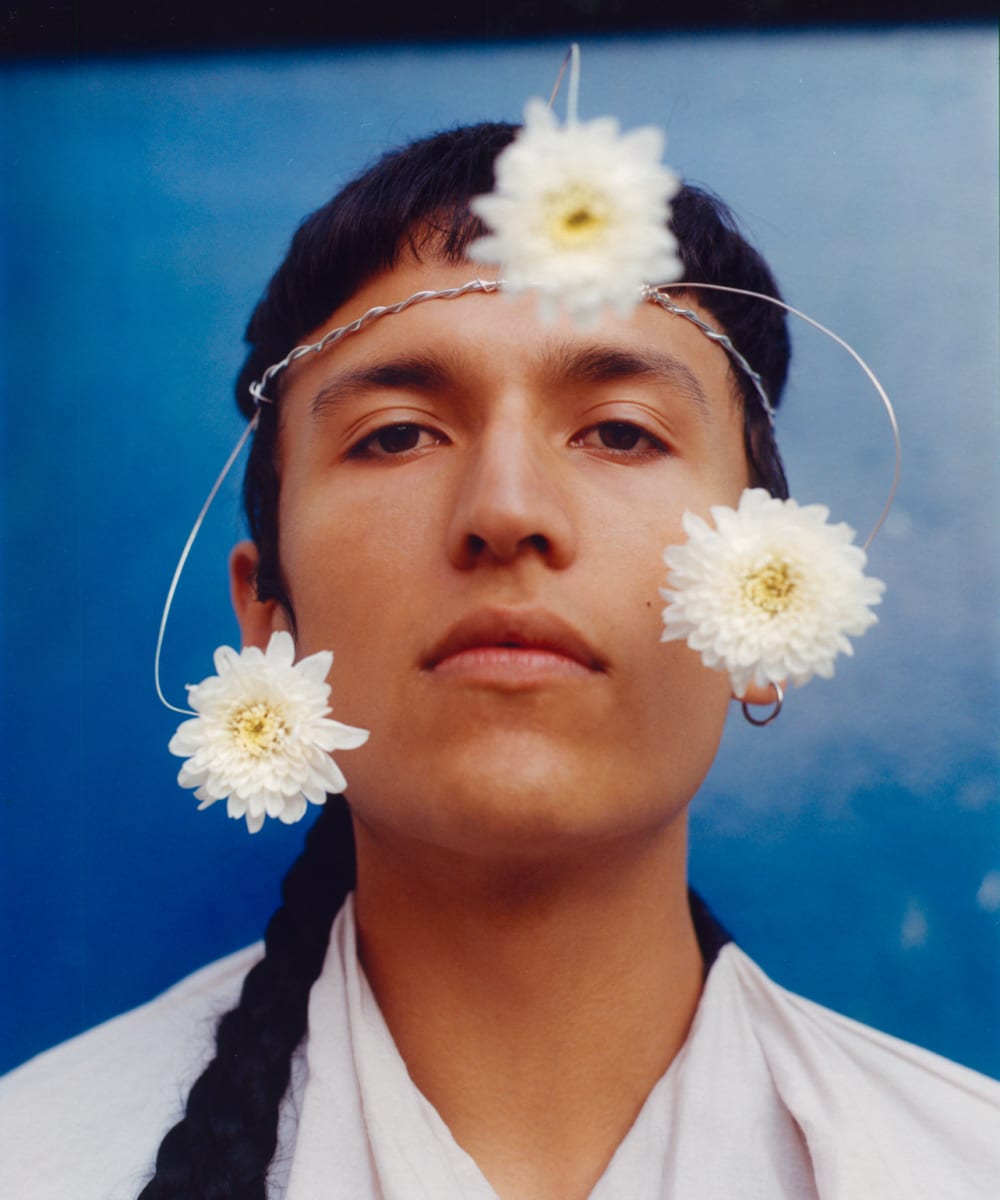 A face in close-up with a long plait to the side, styled with white flowers on a wire around their face and they are wearing a white v-necked garment, just visible