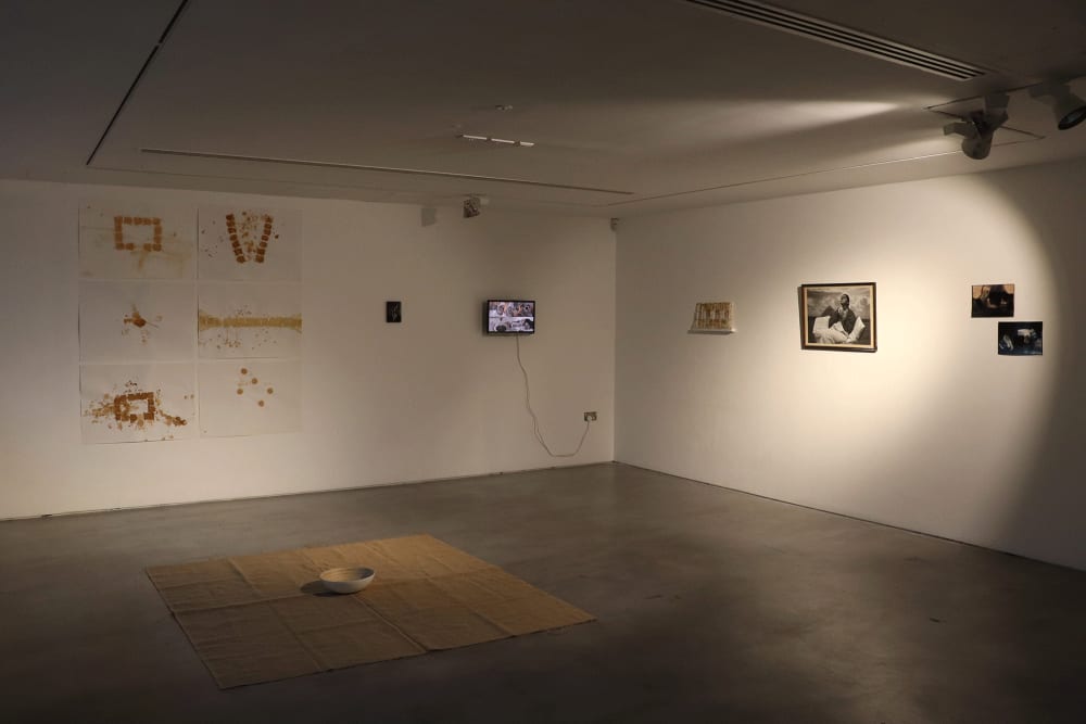 Image shows a large open room with dim over head lighting and artworks hanging on the walls, including a TV screen. On the floor, there is a rug with a ceramic bowl on top.