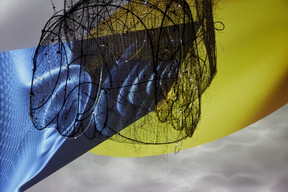 A ceiling display of tangled metal and electrical wires wrapped around a metal cage. Behind the display is a screen displaying waves and a solid yellow background.