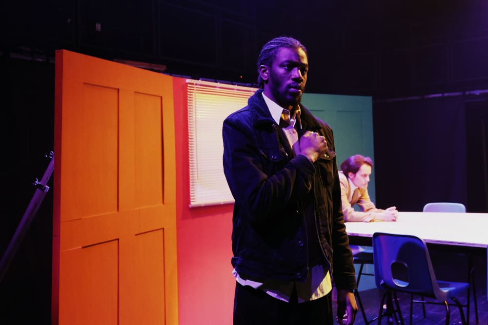 Image shows a man standing with his fist on his heart. In the background are multicoloured doors and another person can be seen leaning over a table. 