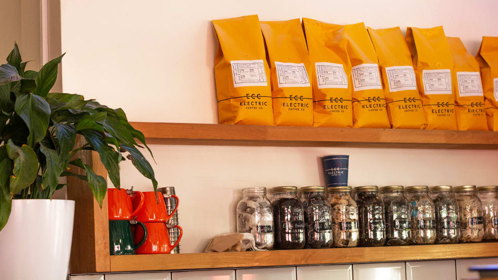 A wooden shelf displays yellow bags of coffee, jars of herbal teas and orange teapots