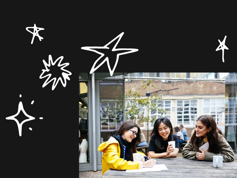 Students sitting outside at a wooden picnic table with coffees and a notebook.