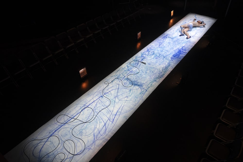 Image shows a long white piece of paper with blue drawings stretched out on a black stage. A person with few items of clothing can be seen lying at the end of the paper.
