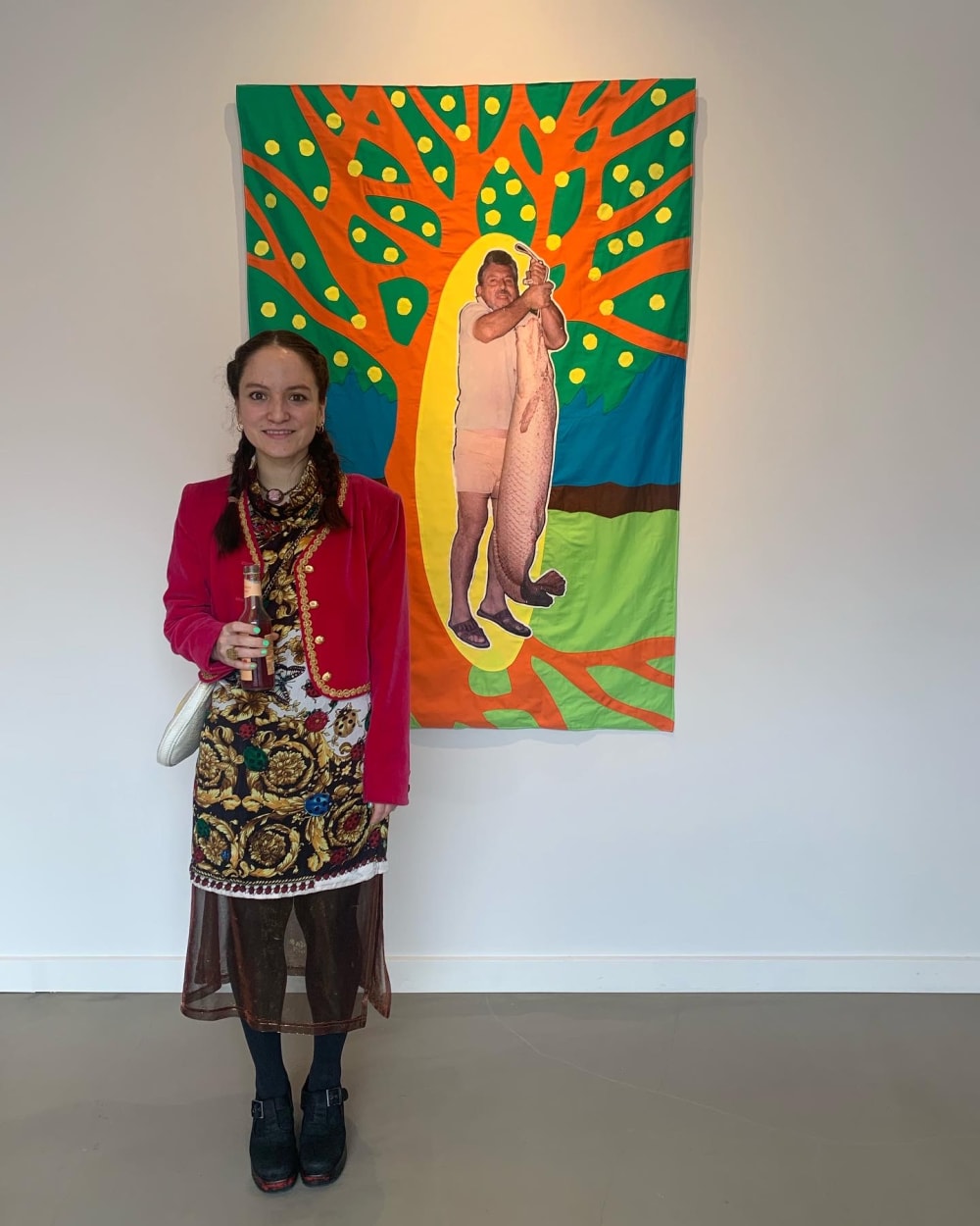 Berenike is standing next to one of her pieces, 'La selva cotidiana, 2022'. An impressionist print of a tree is overlaid with a photo of a man holding a huge fish upside down. 