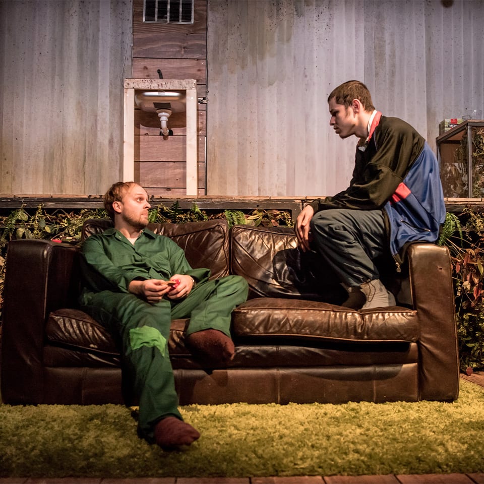 Two actors sit on a sofa, one on the left seat and one on the right arm. Behind the sofa is a wall of foliage.