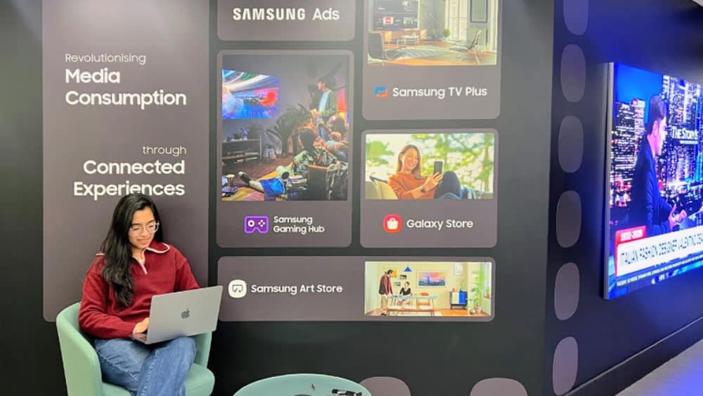 Student sat in office chair in front of digital screen with Samsung creative assets. Student holding laptop.
