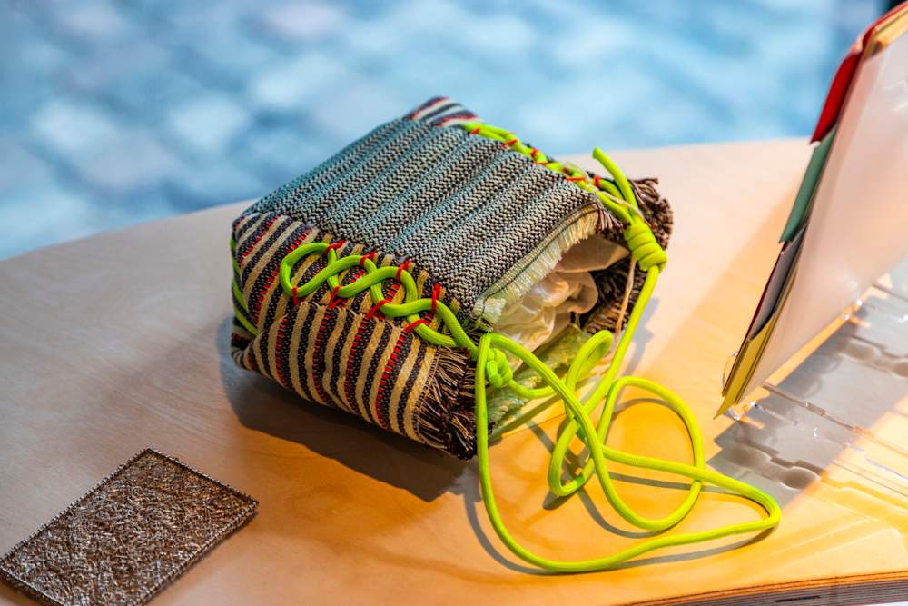 A silver rectangular jewellery item lays on a tabletop next to a small pouch with coloured stripes on the sides and a neon green thin strap stitched down the sides.