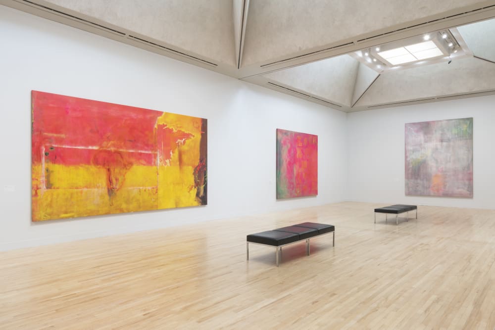 A shot of one of the room at Tate Britain with three abstract pantings by Frank Bowling hanging in it. One is large, red and yellow. One is smaller and red and green. One is pale purple and pink. Also in the otherwise empty room are two benches.