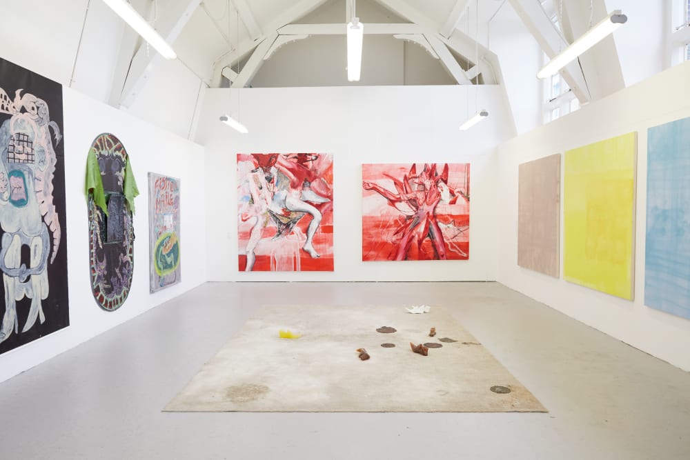 A room of work by BA Fine Art Painting students. In the centre of the room is a floor piece with casts of shoes scattered on a square of pale carpet. On the left wall are three canvases, the first black with a large figure in pastel colours, the second dark and oval with green fabric draped from the top and the third pale with red text on it. On the back wall are two red paintings depicting abstracted figures. To the right are three canvases washed with single colours (l-r) lilac, yellow and pale blue.