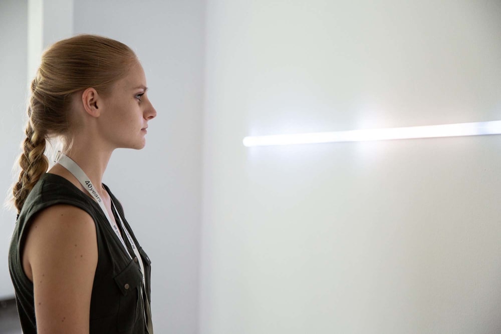A woman in a white room looks towards a white neon tube light.