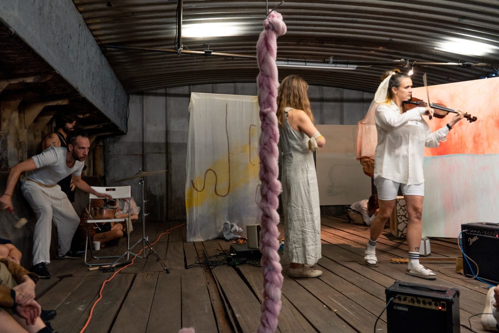 A performance takes place in a dark room with a wooden floor and an industrial curved ceiling. Three performaers including a violin player stand amongst an installation of mixed-media artworks including painted fabric and woven ropes which are suspended from the roof.
