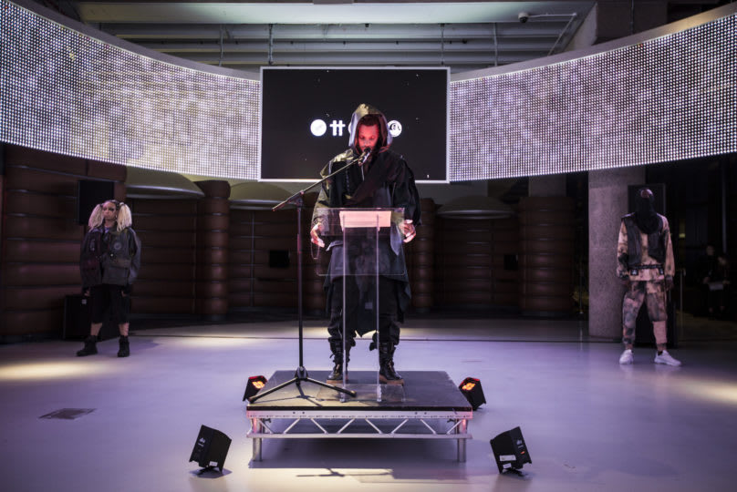 three models in the middle of a performance, one on a staged podium, the other two on either side in the distance. a black screen lit with white text is in the distance. floor lights surround the podium.