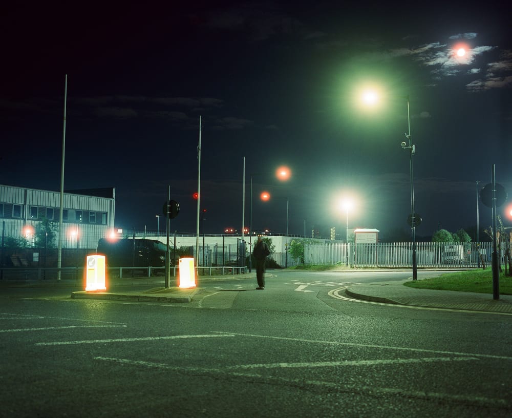 A dimly lit figure is facing the camera, from a distance, standing the centre of the road at night time