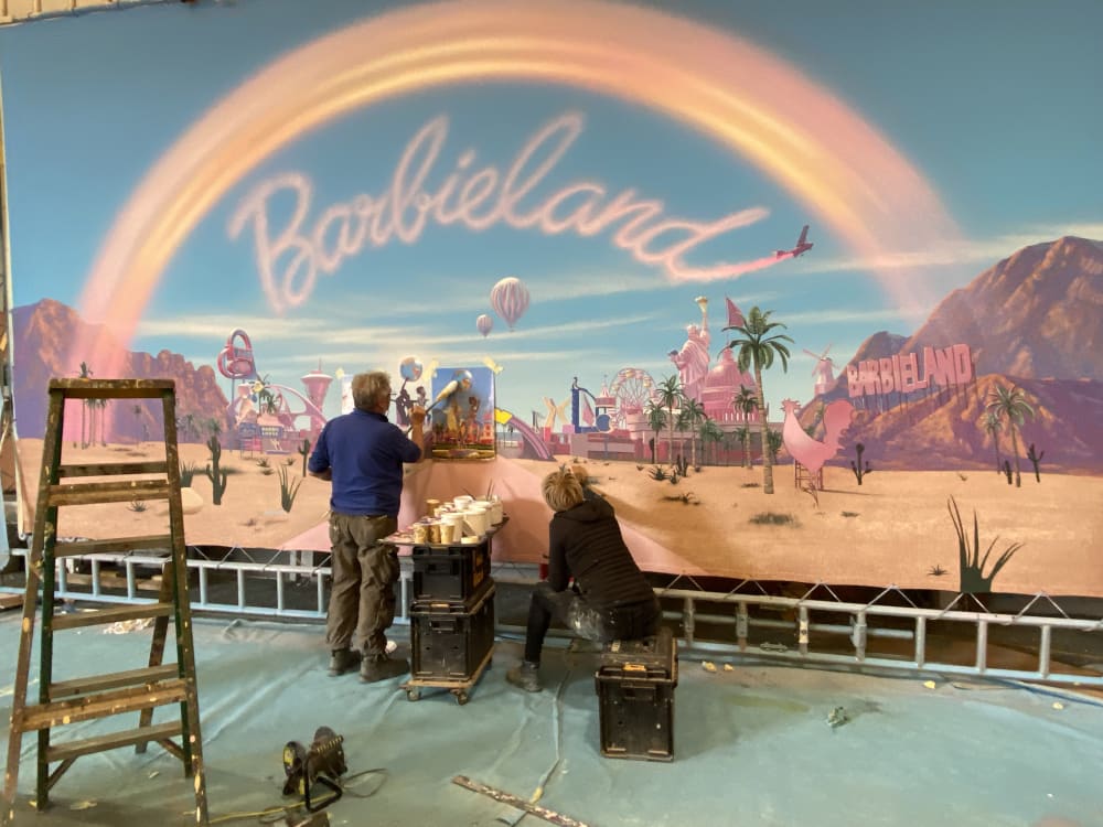 Image shows 2 people painting a set piece for Barbie. The painting is of a pink road with cacti on either side. A colourful pink rainbow runs across the image and the words ‘Barbie Land’ is written under the rainbow. Pink buildings can be seen in the background as if the car is driving away from Barbie Land.