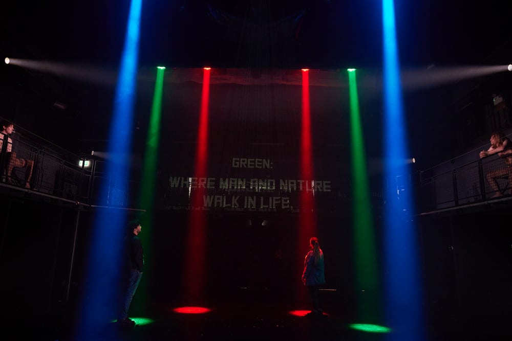 Image shows a dark stage with red blue and green spotlights down either side. 2 people can be seen sitting in the top right and left corners, above the stage. 2 other people are can be seen standing in spotlights on either side of the stage.