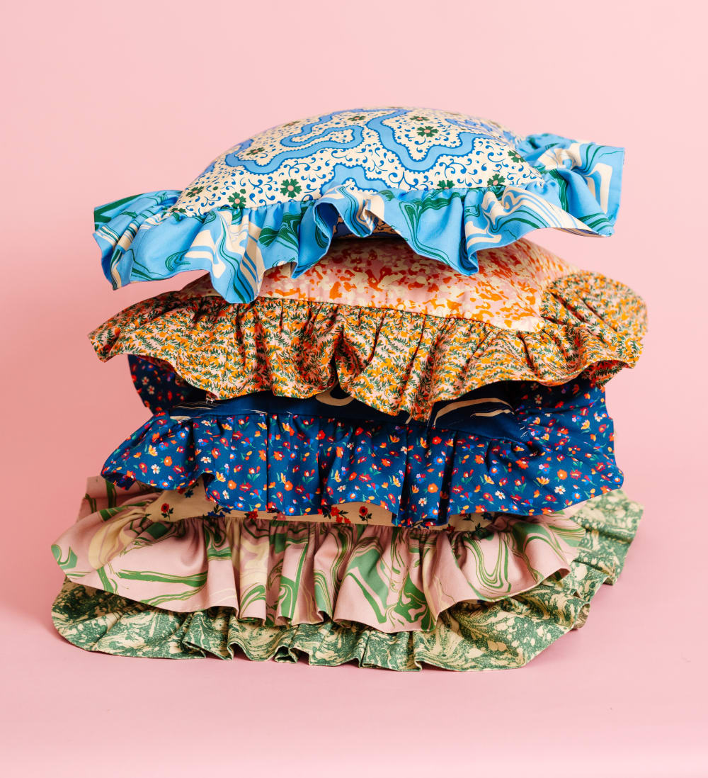 5 cushions piled on top of each other against a pink background. The cushions are all square with a large frill. They each have a different pattern. 