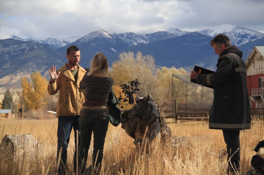 On set in Montana. A man and a woman are standing looking at each other. They're stood amongst grass with trees and mountains in the background. To the right, is a man looking at a clipboard. 