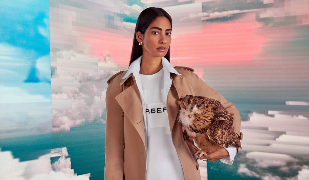 Person wearing a brown trenchcoat and white Burberry tshirt, holding an owl, standing in front of a digitally created sky background.